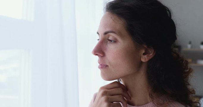 Pensive worried young adult woman looking outside through window. Thoughtful serious lonely lady feeling sad or melancholic, reflecting alone, thinking or loneliness, solitude concept. Close up view