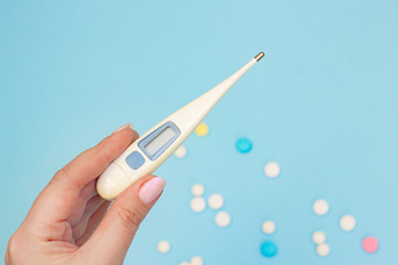 Electronic thermometer in hand on a blue background with scattered pills. Elevated body temperature as a symptom of coronavirus infection