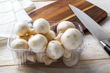 Heap of whole raw champignons in a plastic food tray, cutting board and knife on a wooden table. Mushrooms as vegetable protein, raw food diet, vegetarianism.