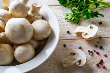 Heap of whole raw fresh champignons in a white bowl, bunch of parsley and pepper on a wooden table. Mushrooms as vegetable protein, raw food diet, veganism.
