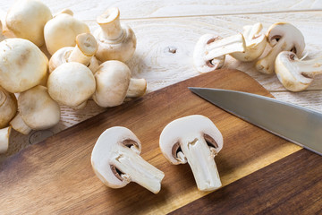 Two halves of champignon and knife on a cutting board near heap of raw fresh mushrooms on a wooden table. Mushrooms as vegetable protein, raw food diet.