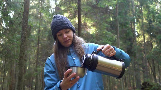  Girl opens a thermos and drinks hot tea from thermos sitting on the tree in the forest. 4K