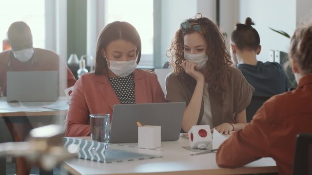 Two female colleagues in protective face masks sitting together at desk and discussing project on laptop while working in office during Covid-19 outbreak