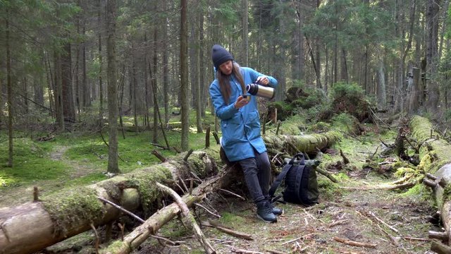  Girl opens a thermos and drinks hot tea from thermos sitting on the tree in the forest. 4K