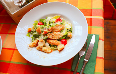 Caesar salad with croutons, quail eggs, cherry tomatoes and grilled chicken in a white plate on a table in a restaurant. Flatlay. Top down. 