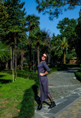 Nice, brunette woman in a dress walks in the street. Enjoys a sunny spring day. Girl traveling in Batumi, Georgia. Green park at background.
