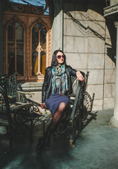 Obraz premium Fashion, brunette woman in a dress and leather black jacket sitting on old iron chair outdoors. Girl model posingl in Batumi, Georgia. Old city at background.