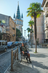 Obraz premium Brunette woman with a positive smile in a dress and leather black jacket walks the street and enjoys a sunny spring day. Girl travel in Batumi, Georgia. Old city at background.