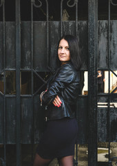 Brunette hipster woman with a positive smile in a leather black jacket posing in the city. Girl model in the Batumi, Georgia. Gates at the background.