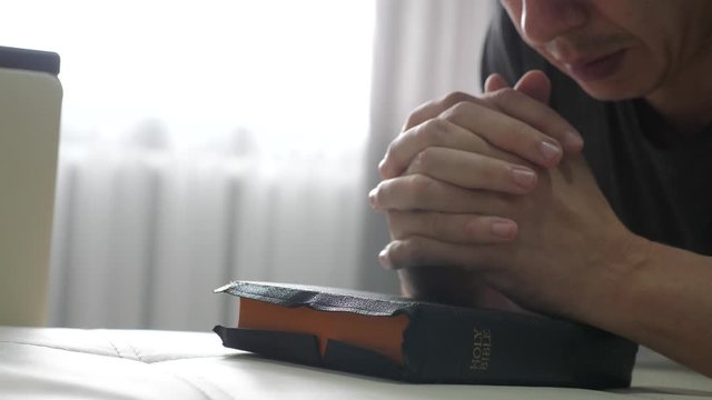 man praying indoors at bedtime on bible. lifestyle religion concept evening prayer human brunette hands on bible praying by bed