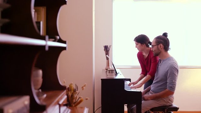 Panoramic side view of man practicing musical lessons indoors. House lifestyle learning music. Piano lessons at home. Female teacher giving a keyboard piano class to student. Couple playing the piano.