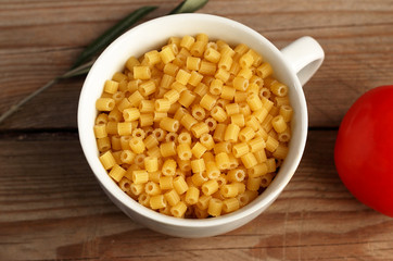 Tazza bianca riempita con pasta italiana cruda su fondale di legno grezzo.