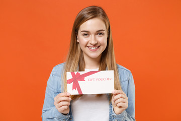 Fototapeta na wymiar Smiling young woman girl in casual denim clothes posing isolated on bright orange wall background studio portrait. People sincere emotions lifestyle concept. Mock up copy space. Hold gift certificate.