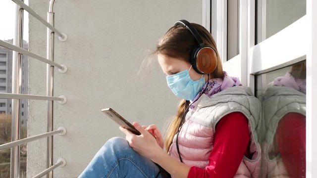 teenager girl in mask and headphones, uses smartphone, on open balcony. spring sunny day. concept of quarantine, chat online. stay at home. coronavirus epidemic.