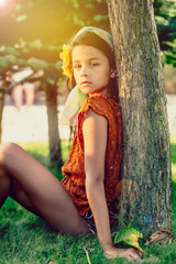 Beautiful little girl brunette with long hair and a hairpin in the form of a flower on her head sitting near a tree in the park. Summer vacation concept.