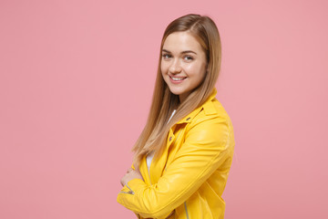 Side view of smiling young woman girl in yellow leather jacket posing isolated on pastel pink background in studio. People lifestyle concept. Mock up copy space. Holding hands crossed, looking camera.