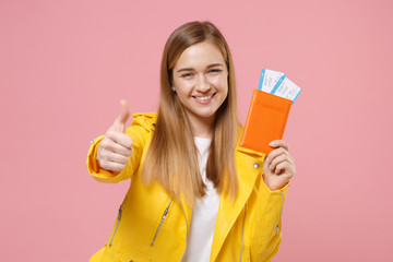 Smiling young woman girl in yellow leather jacket posing isolated on pastel pink wall background. People lifestyle concept. Mock up copy space. Hold passport tickets boarding pass, showing thumb up.