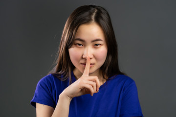 Smiling asian woman with long hair holding finger on her lips over grey background. Gesture of shhh, secret, silence. Close up.