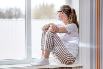 Home quarantine. Caucasian woman on sick leave is sitting at window in a medical protective mask, looking sadly at street. Poor health, a seasonal epidemic of flu virus and colds. Home treatment.