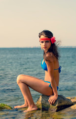 Travel Concept. Girl tourist sits on a mountain stone. Girl sitting on a cliff face over the sea. Clean sea and wild seascape.Young brunette girl with flower on her head enjoying vacation.Copy space