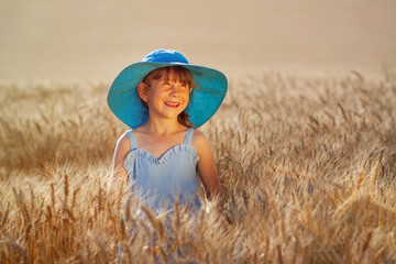 little red-haired girl in blue dress and blue round hat stands happily in gold field of wheat. Summer concept