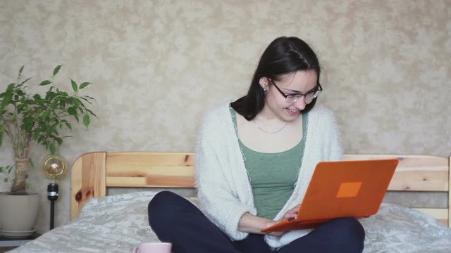 Young attractive girl is actively browsing the news feed on a laptop while sitting on a bed at home. Young Cute girl is working on a laptop while sitting on a bed. Working at home.