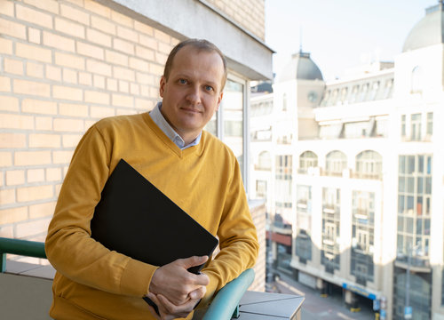 Middle Aged Man Working From Home On Laptop And Having A Break On Balcony With Nice City View.