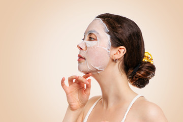 Portrait of a young woman with a fabric moisturizing mask on her face which gracefully emphasizes the chin with her hand. Beige background.Copy space. The concept of beauty, lifting and self-care