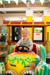 A very joyful and happy little blond boy of three years old is riding in a yellow car with a green...