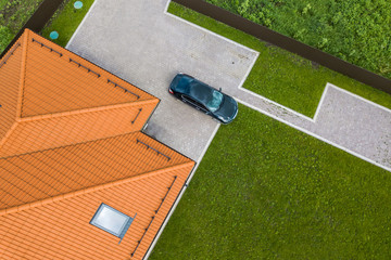 Aerial top view of house shingle roof with attic windows and black car on paved yard with green...