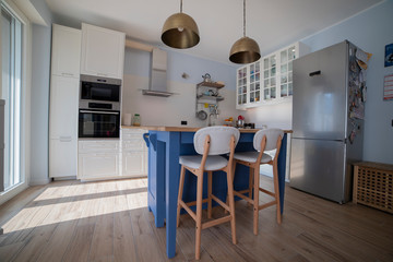White, bright kitchen with blue island and double brass pendant lights. Low view.