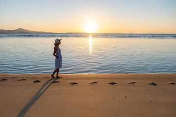 Young woman in straw hat and a dress walking alone on empty sand beach at sunset sea shore. Lonely...