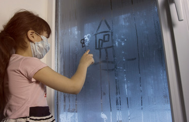 A little girl in a medical mask sits on the windowsill and draws a house on the fogged window. Quarantine, stay at home. The who's recommendation