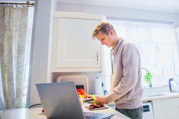Young caucasian man working from home. Freelancer cooking vegetarian lunch on the kitchen table and working on pc laptop. Home office, remote workplace concept. Soft selective focus. Copy space.
