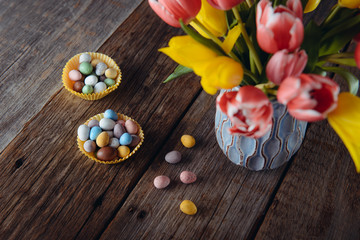 Obraz na płótnie Canvas Top view two paper bags with Easter candies covered eggs in various pastel colors near vase with fresh tulips on rustic wooden table. Fastive holiday sweets. Copy space.