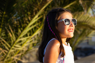 Happy little girl with colorful flowers frame sunglasses looking at sun. Palm trees reflecting on eyeglasses of smiling kid on summer vacation. Sunscreen, ultraviolet light, eyes protection concepts