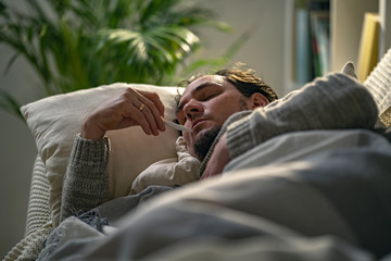 Sick men lying on a sofa and putting his hand on head, holding thermometer.