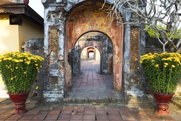 Fototapeta na wymiar Ancient Corridor with Weathered Brick Walls and Distant Portals with Yellow Flowers in a Flowerpot. Hue, Vietnam Southeast Asia