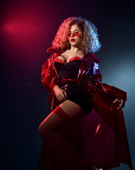 Blonde curly female in sunglasses, red and black lace sexy lingerie, stockings, transparent coat. She posing on gray background