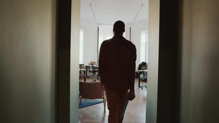 Rear view following shot of African American man in casual outfit holding clipboard while walking through hallway and entering open space office