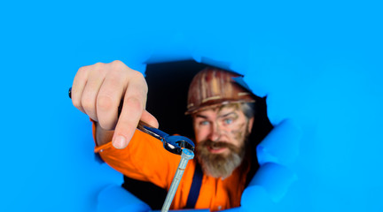 Through paper worker in protective helmet. Builder holds wrenches. Repairing tools. Spanner. Bearded builder man with wrenches looking through paper. Man builder with wrenches looking through paper.