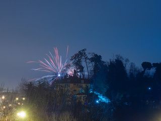 Bright flash of fireworks near the roof of a yellow house on a background of blue sky and trees