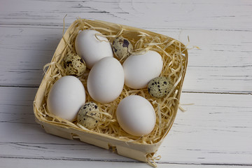 white large chicken and quail eggs in a basket with straw on a white light wooden background