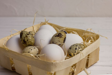 white large chicken and quail eggs in a basket with straw on a white light wooden background