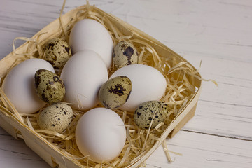white large chicken and quail eggs in a basket with straw on a white light wooden background