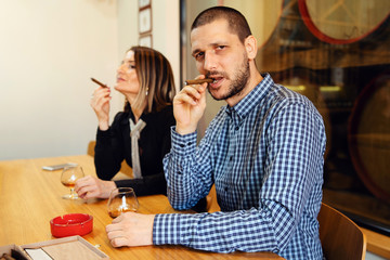 Side view of adult caucasian man and woman couple sitting by the table smoking tobacco cigar and drinking cognac brandy looking to the camera in day wearing shirt
