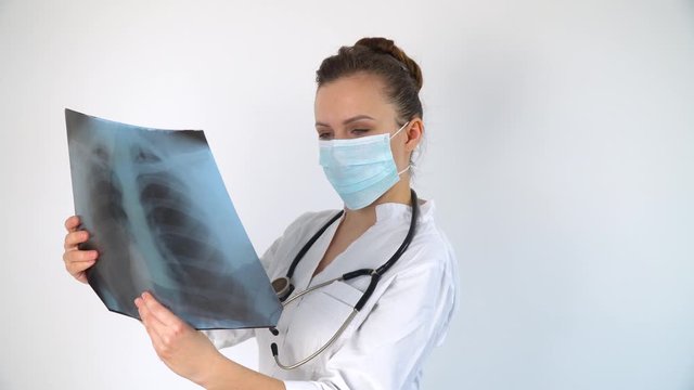 Young caucasian doctor in protective mask examine x-ray of lungs, holding it in hands indoors. Specialist hold transparent image of chest in arms, and carefully researches it for coronavirus