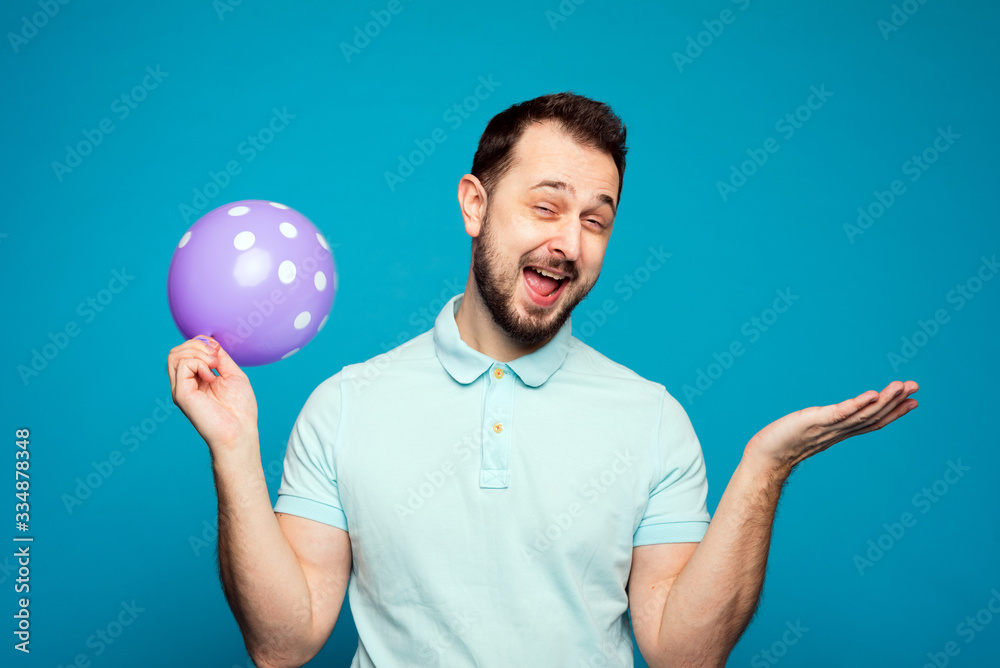 Wall mural A handsome man holds a balloon in his hand and rejoices and screams at a holiday or birthday. A man inhaled laughing gas, helium and laughs on a blue background in the studio