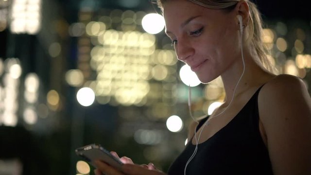 Young Caucasian hipster girl searching chapter of audio book in smartphone application during evening time on urban setting,slow motion of millennial woman listening song in electronic earphones