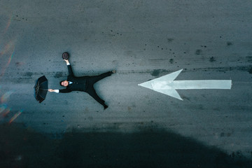 Man in black suit holding a black umbrella is being blow into the air on a street with an arrow